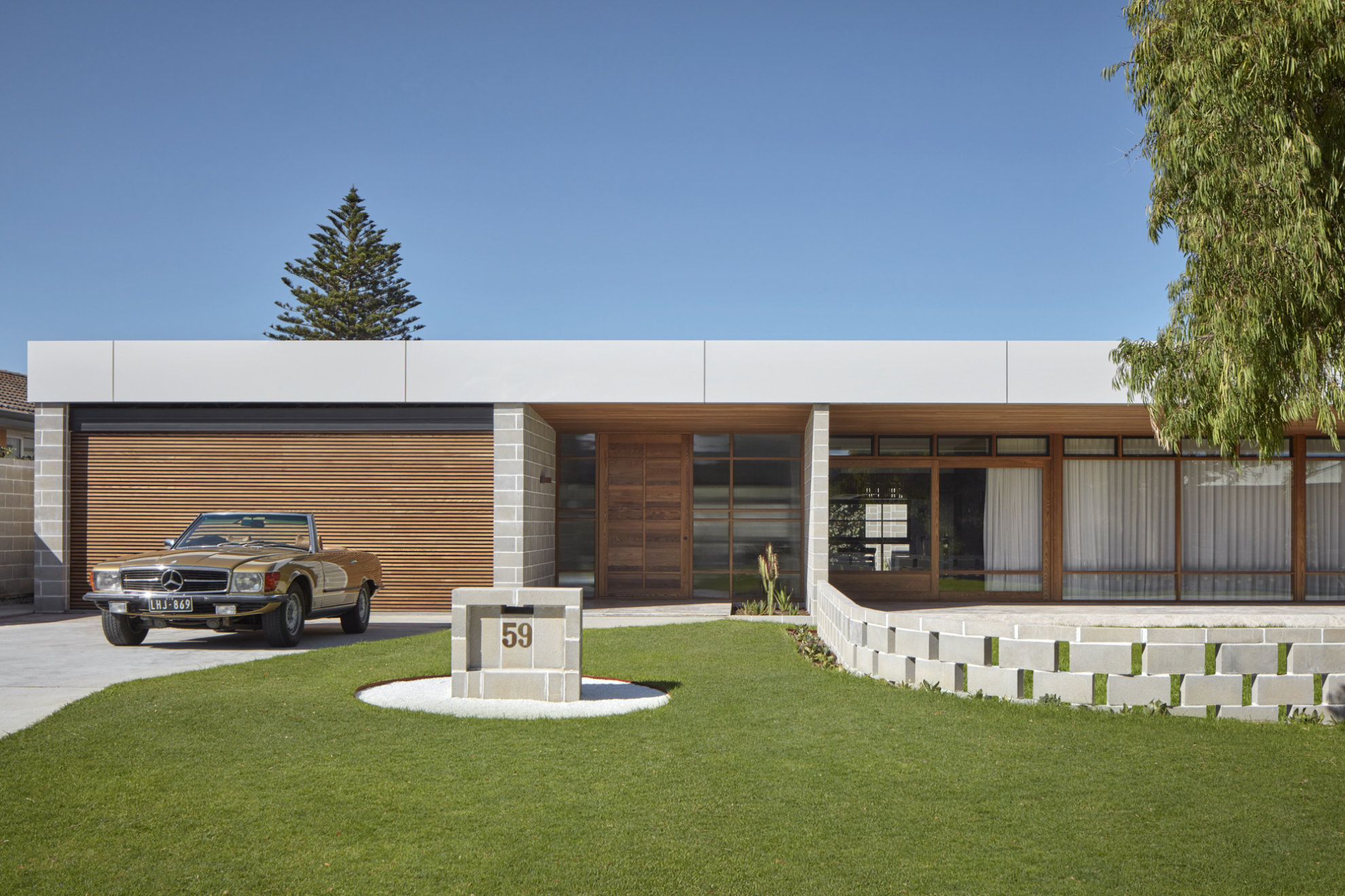 The house’s wide, rectangular, single storey façade: The wooden, glass, and concrete brick exterior stretches across the image. A short, alternating cement block divider extends from the house and curves around the house. The house number: 59, is displayed on a concrete block postbox on the lawn. A vintage light brown Benz convertible is parked in the driveway, in front of the custom, wooden-slatt garage door.