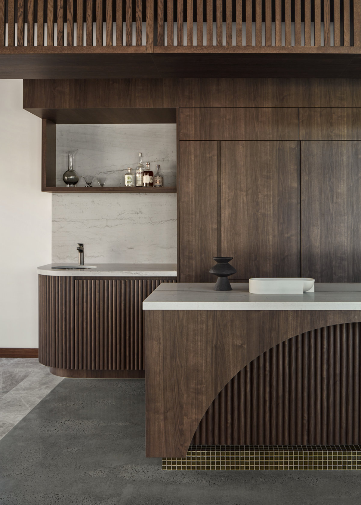 Kitchen interior with dark wood paneling and green tile highlights. The kitchen island countertop and the sink area backsplash are in white marble. A black sculptural vase and a white oval tray is on top of the island.