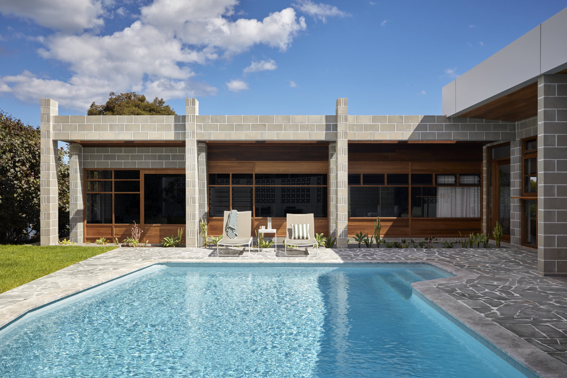 A sunlit pool in front of a single-storey Modernist house built with large concrete blocks. The entire visible facade of the house is lined with large glass windows framed with wood.