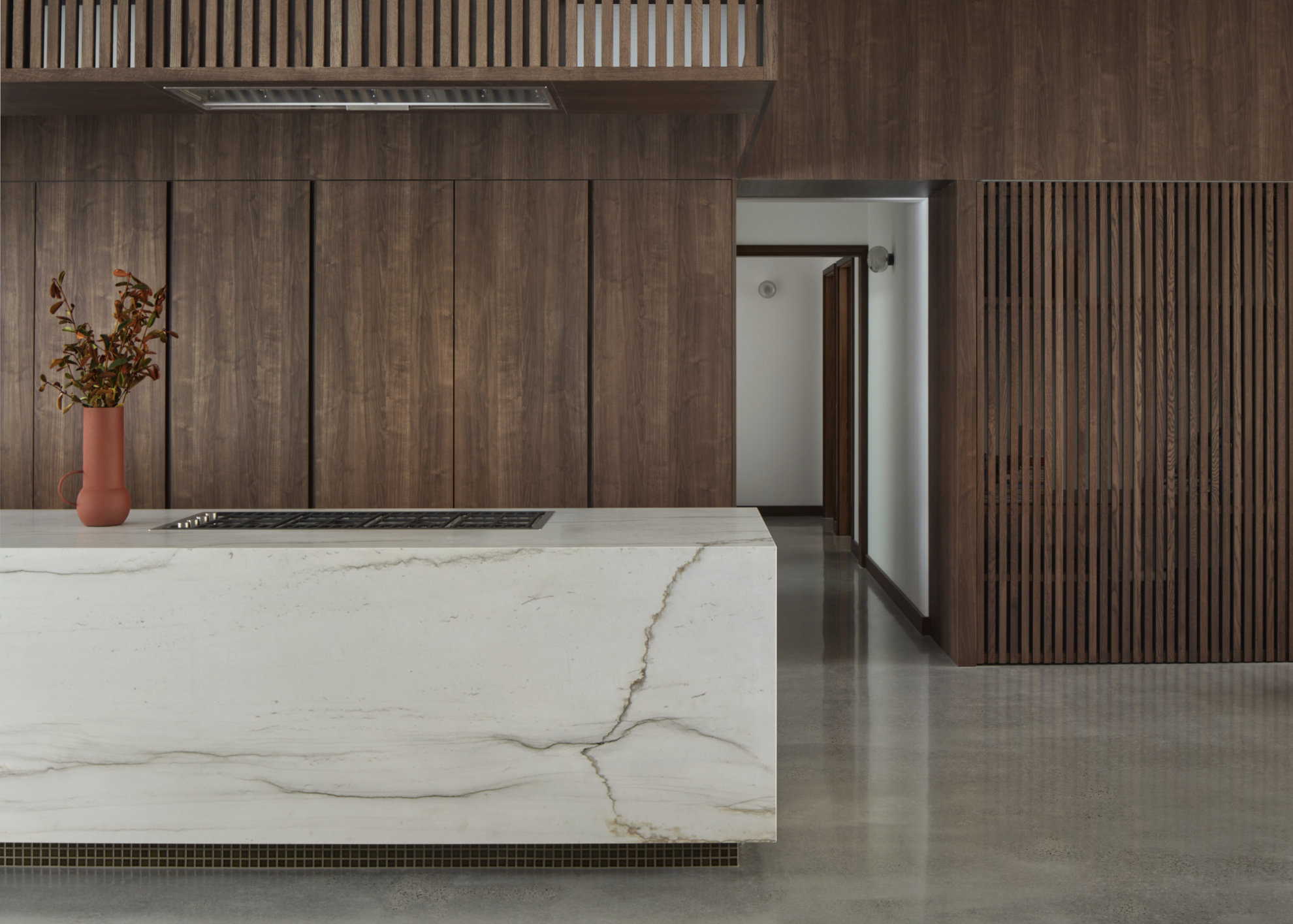 A stone kitchen bench in front of timber panelling with hidden cabinetry. A stovetop sits flush with the surface of the bench. The only object on the bench is a red vessel containing reddish foliage.