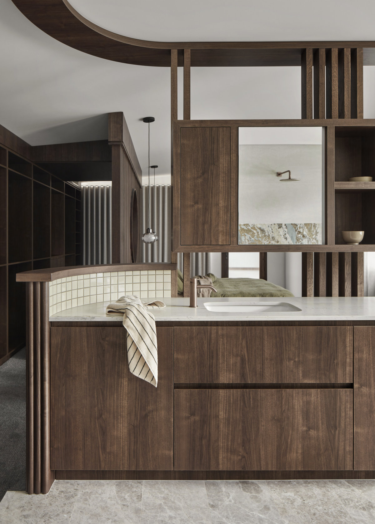 Ensuite with dark wood cabinetry, white marble countertop, brass cylindrical fittings can be seen atop the sink and reflected in the mirror. A small striped towel hangs from the countertop. In the background, above the bed, are two oval shaped pendant lights.