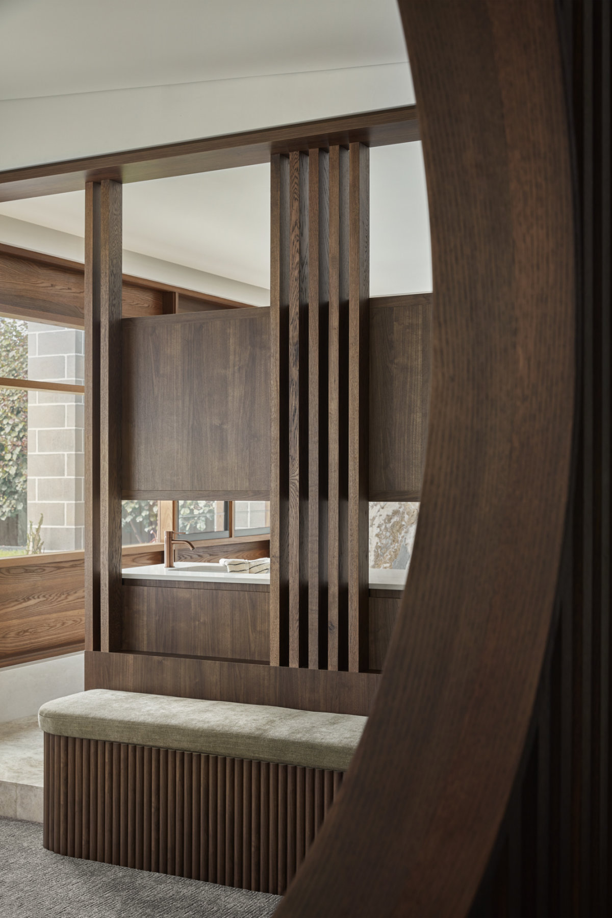 Views in to the ensuite and backyard through a bedroom portal and slats. Attached to the bedroom is a short bench with vertical wooden slats and a olive cushioned top.